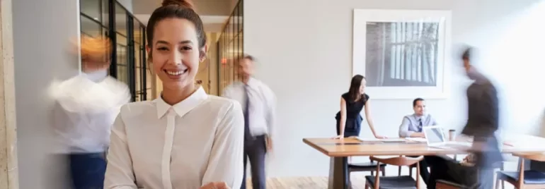 Smiling woman in white shirt stands with blurry office workers in background.